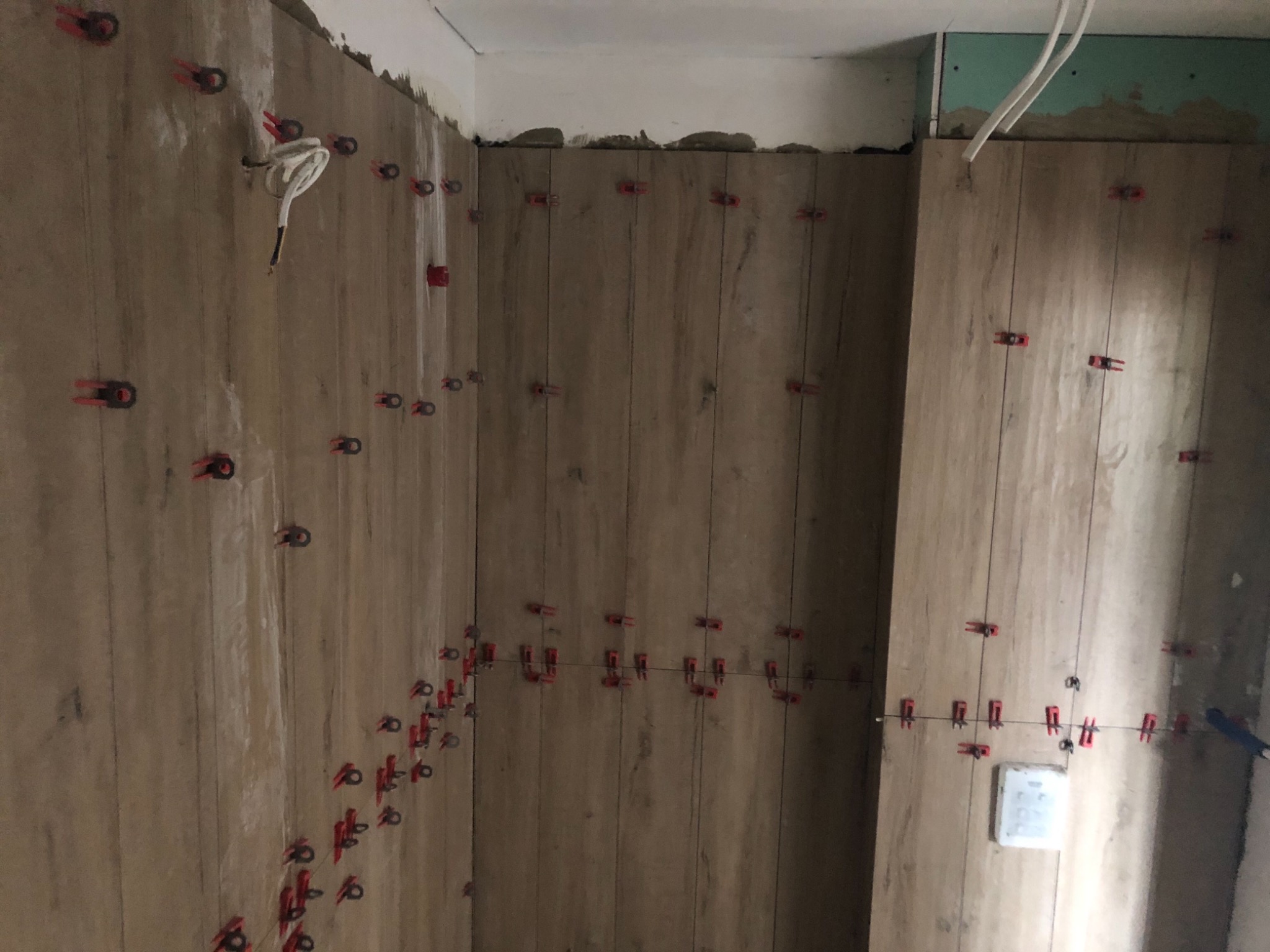 Ściana łazienki w trakcie układania płytek imitujących drewno, z widocznymi czerwonymi klipsami poziomującymi i wystającymi przewodami elektrycznymi.