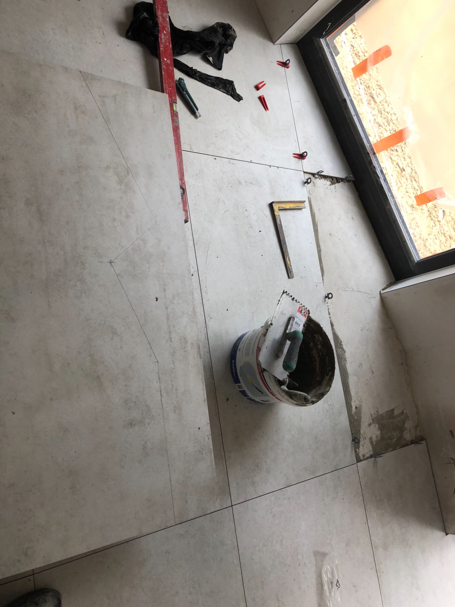 Fragment podłogi z jasnych płytek, w trakcie układania, widoczne narzędzia: poziomica, wiadro z zaprawą, szpachelka, kątownik oraz krzyżyki dystansowe, na płytkach zaznaczone linie cięcia.