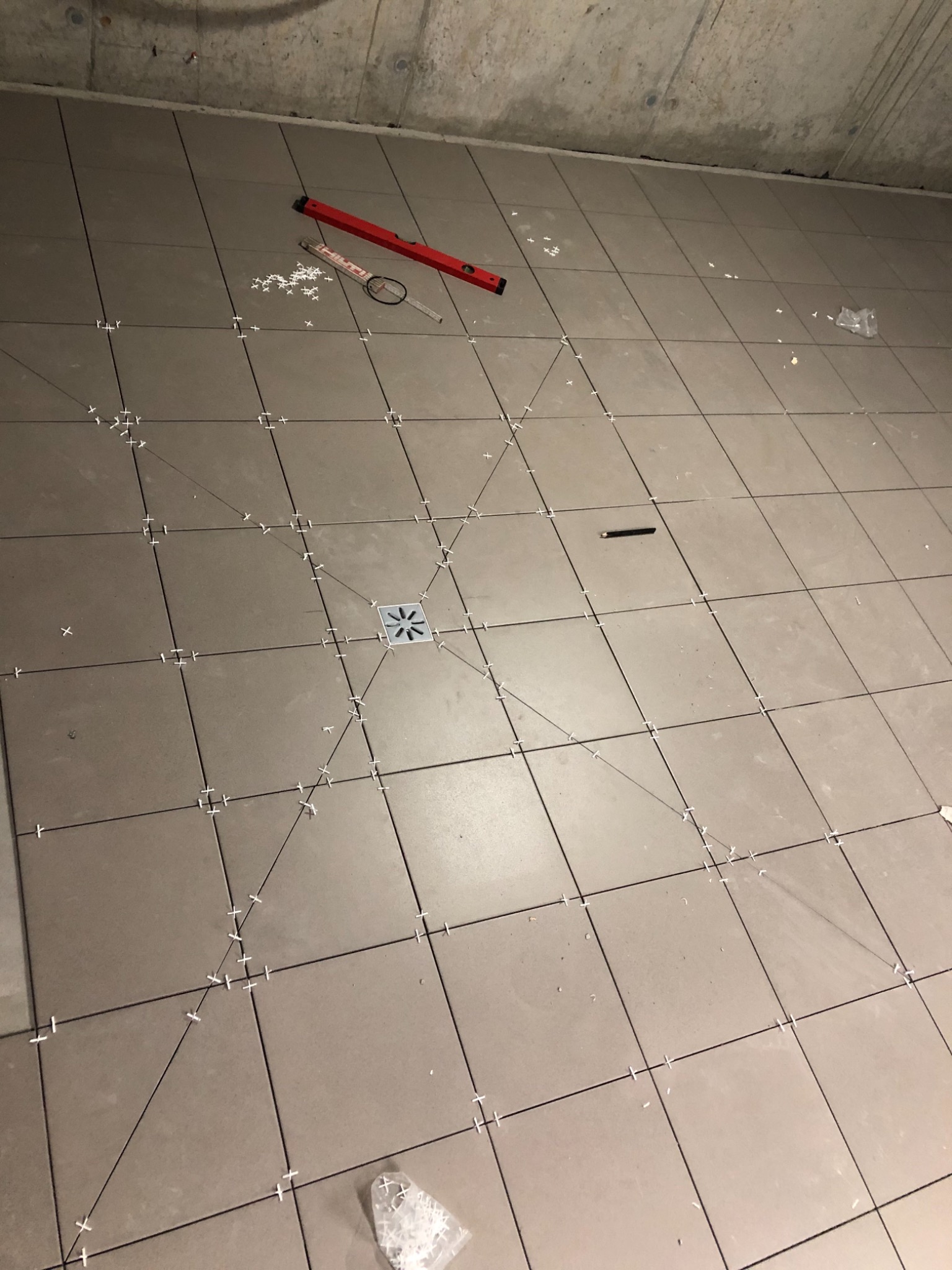 Ułożone szare płytki podłogowe z krzyżykami dystansowymi, widoczny odpływ liniowy, poziomica i ołówek na powierzchni, surowy betonowy sufit w tle.