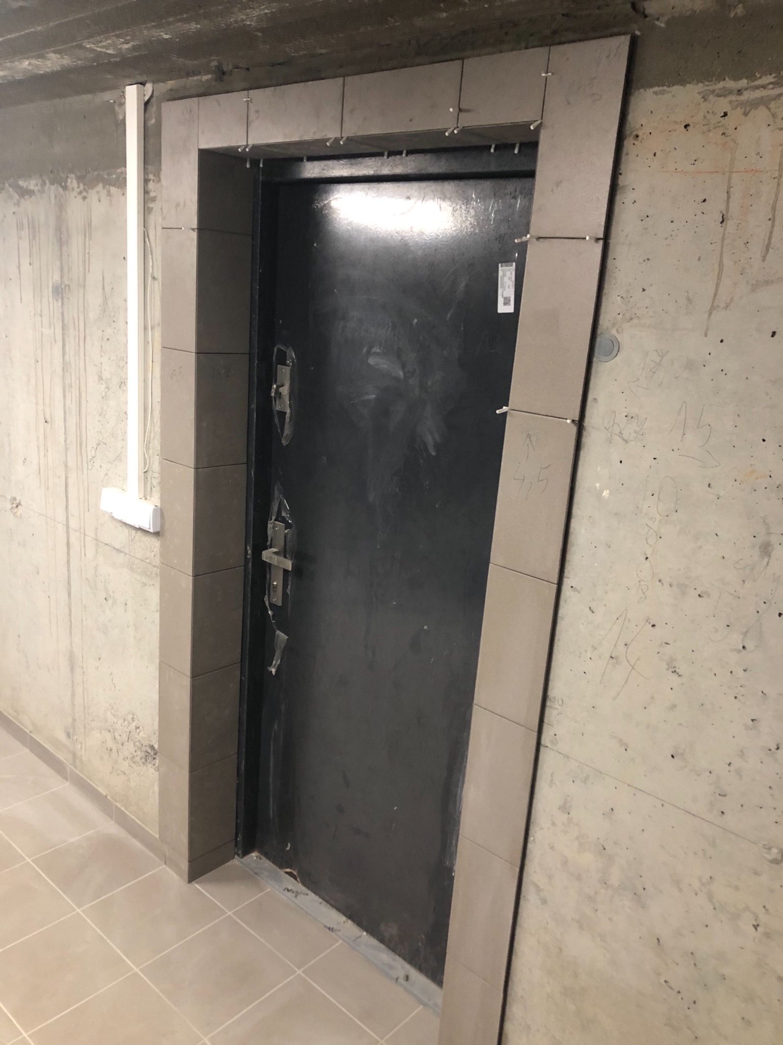 Montaż płytek wokół czarnych drzwi wejściowych w surowym wnętrzu, widoczne krzyżyki dystansowe, ślady po ołówku na ścianie i podłodze, betonowy sufit.