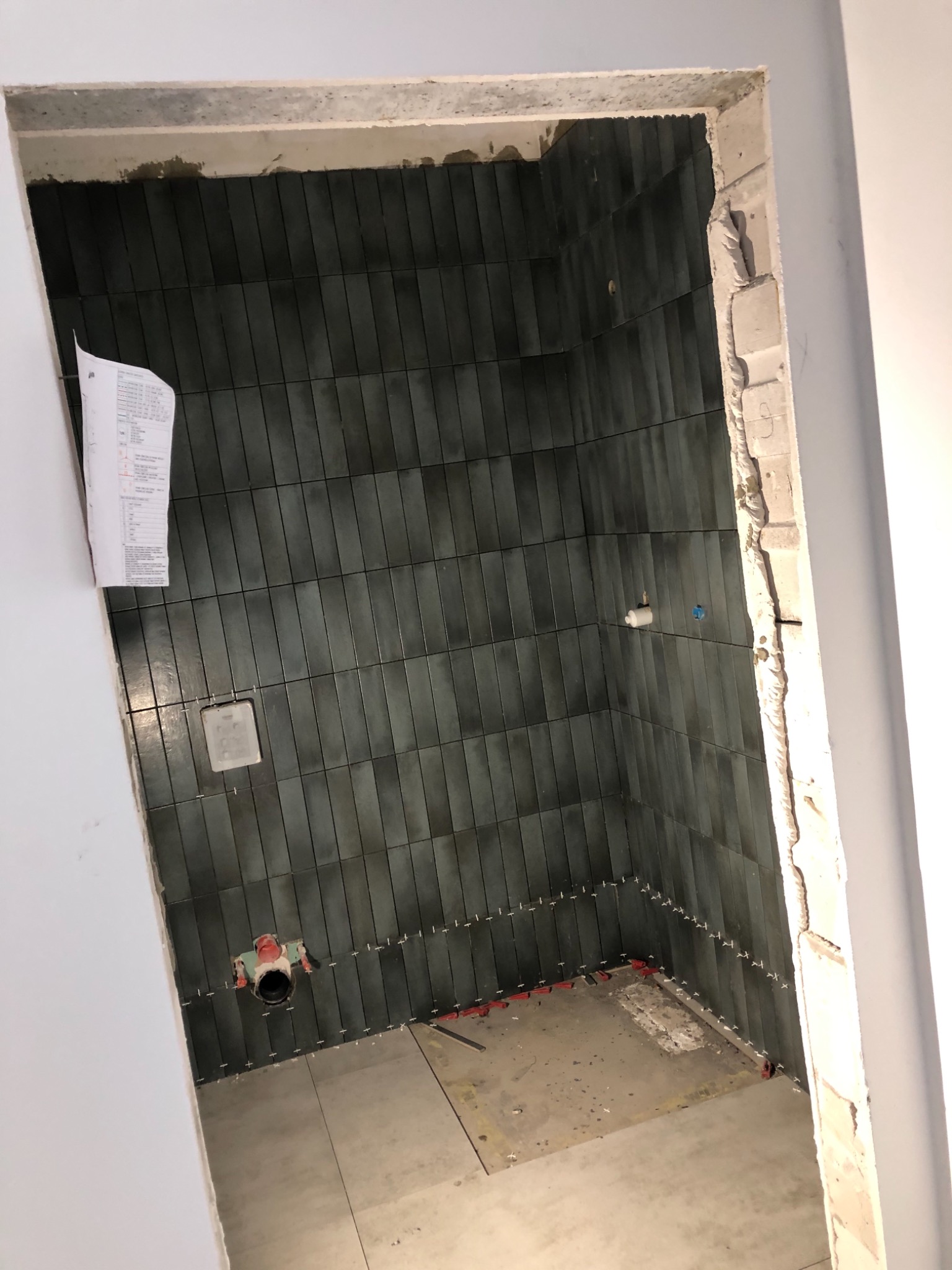 Wnętrze łazienki w trakcie prac wykończeniowych: pionowo ułożone, ciemnozielone płytki na ścianach, widoczne krzyżyki dystansowe i poziomica, surowy beton wokół otworu drzwiowego, kartka...