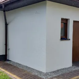 Świeżo otynkowany, biały budynek z brązowymi oknami i drzwiami, widoczne narzędzia i wiadro farby na zewnątrz, sugerujące prace wykończeniowe.