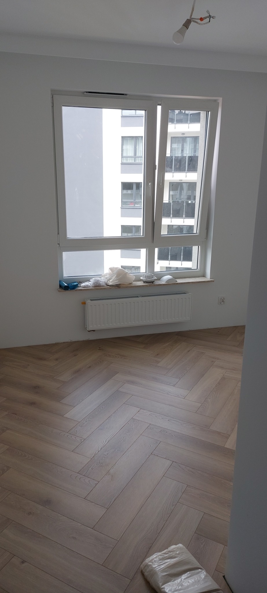 Pokój po remoncie z nową podłogą ułożoną w jodełkę francuską, białe ściany, grzejnik pod oknem z widokiem na budynek mieszkalny, żarówka wisząca na kablu.