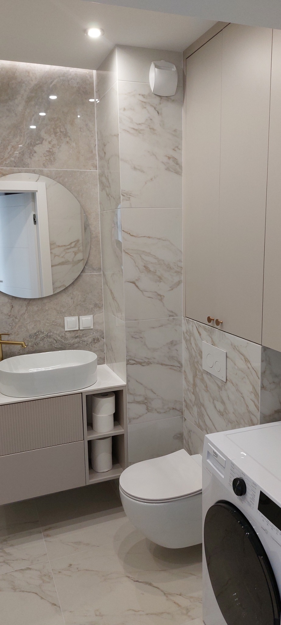 Nowoczesna łazienka wykończona płytkami imitującymi marmur, z owalną umywalką nablatową, podwieszaną toaletą i pralką Beko w rogu.