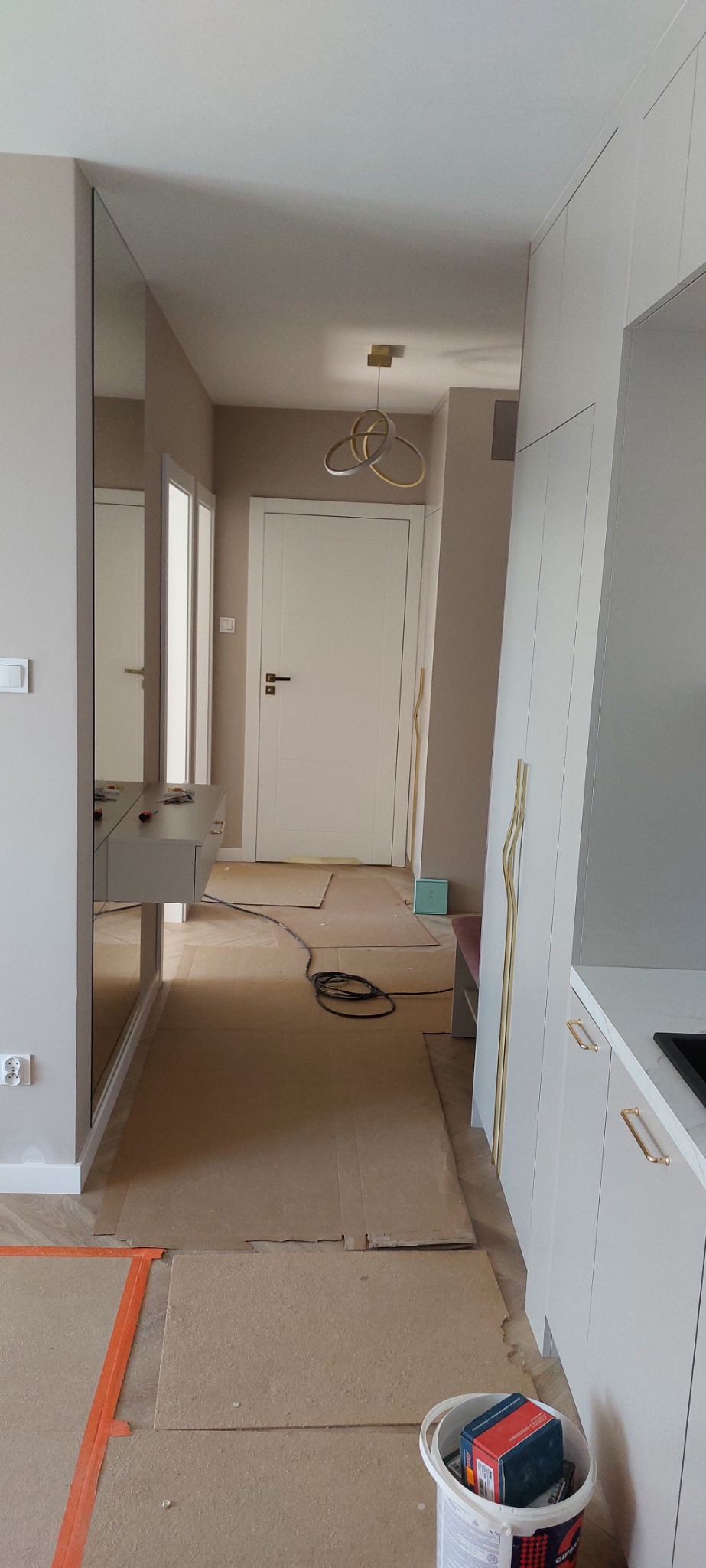 Przedpokój w trakcie prac wykończeniowych: podłoga zabezpieczona kartonem, widoczne narzędzia i materiały, lustro na ścianie, zabudowa meblowa z złotymi uchwytami, nowoczesna lampa sufitowa.