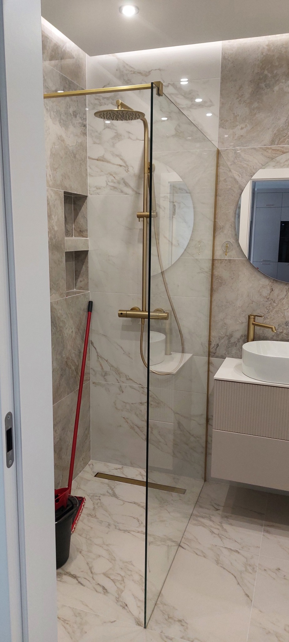 Nowoczesna łazienka z kabiną prysznicową typu walk-in, złotą armaturą i marmurowymi płytkami, widoczny mop w rogu.