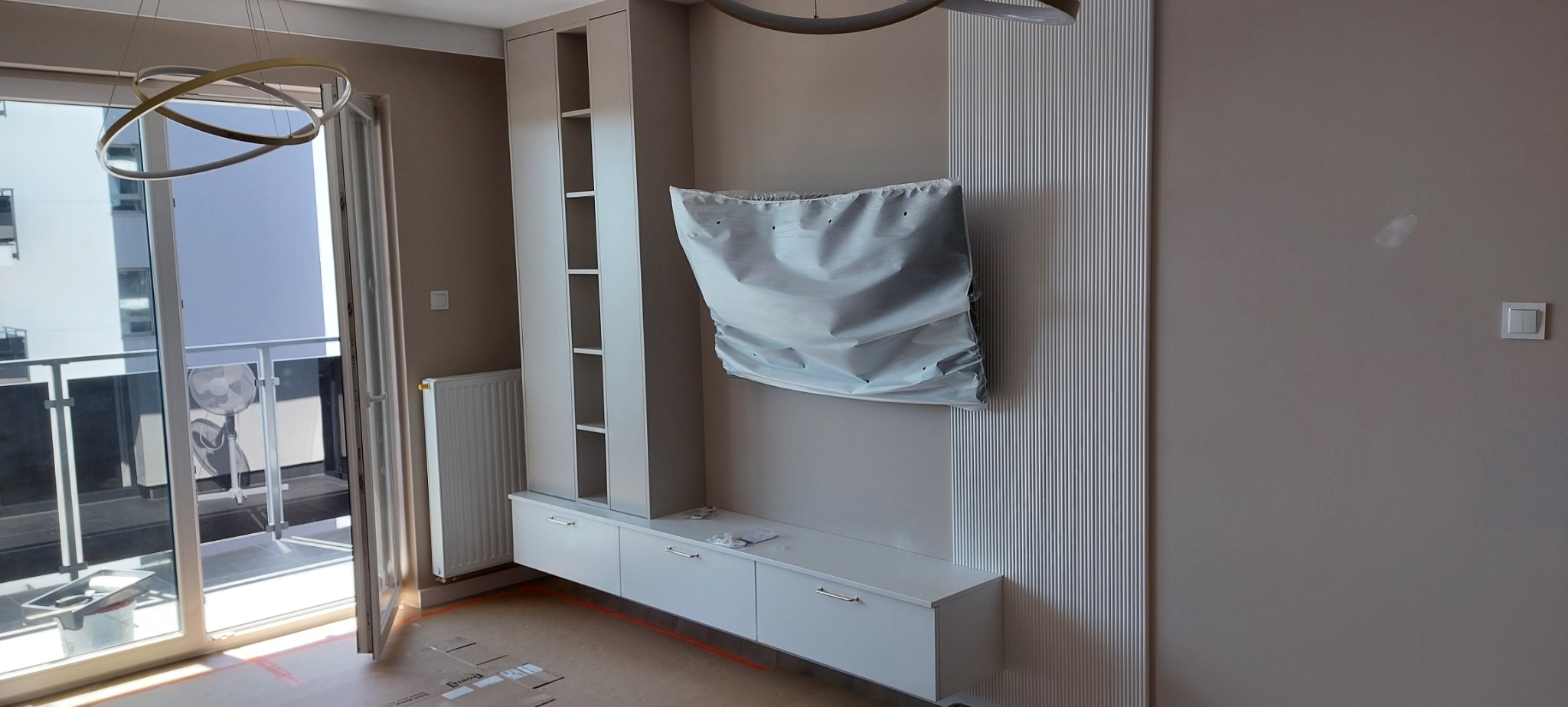 Wnętrze pokoju w trakcie wykańczania, z białą zabudową meblową, pionowymi panelami dekoracyjnymi na ścianie, osłoniętym folią telewizorem i widokiem na balkon.