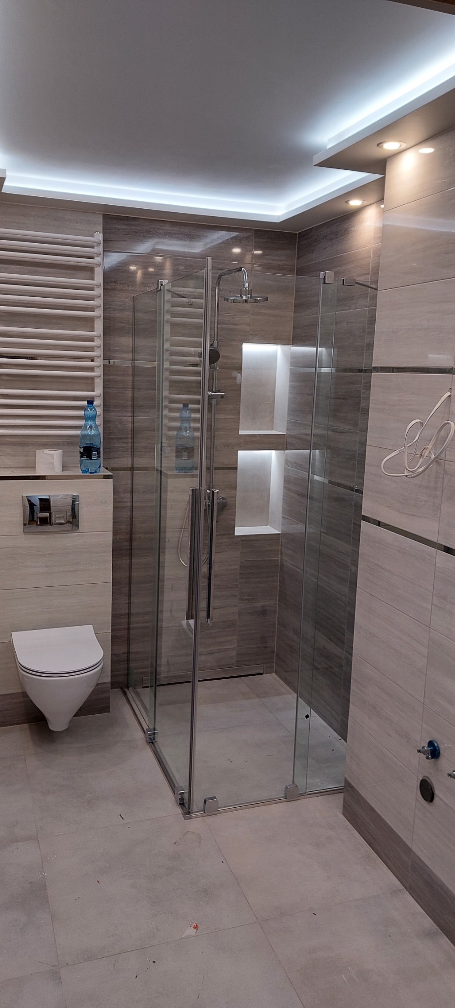 Nowoczesna łazienka w Kikole z kabiną prysznicową typu walk-in, wykończona płytkami imitującymi drewno, z podświetlanymi wnękami i grzejnikiem drabinkowym.