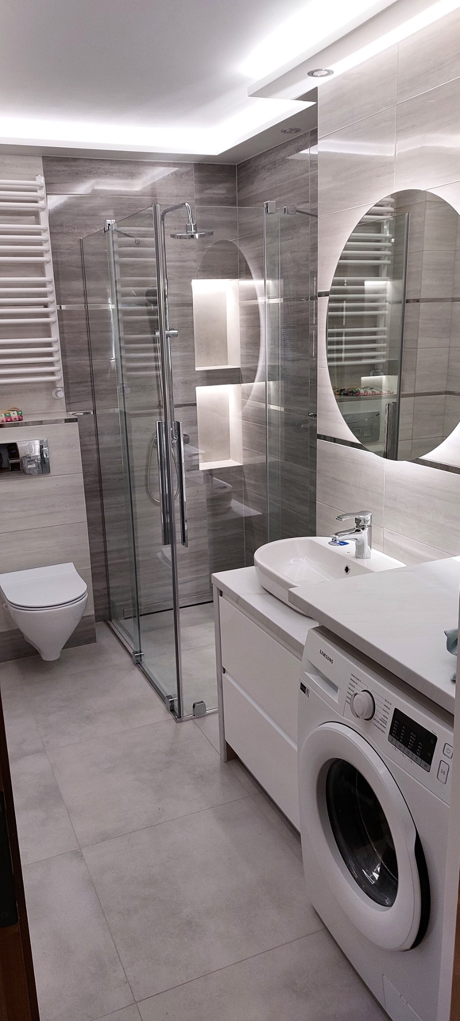 Nowoczesna łazienka w szarościach z kabiną prysznicową, wiszącą toaletą, okrągłym lustrem z podświetleniem LED i pralką Samsung ukrytą pod blatem umywalki.