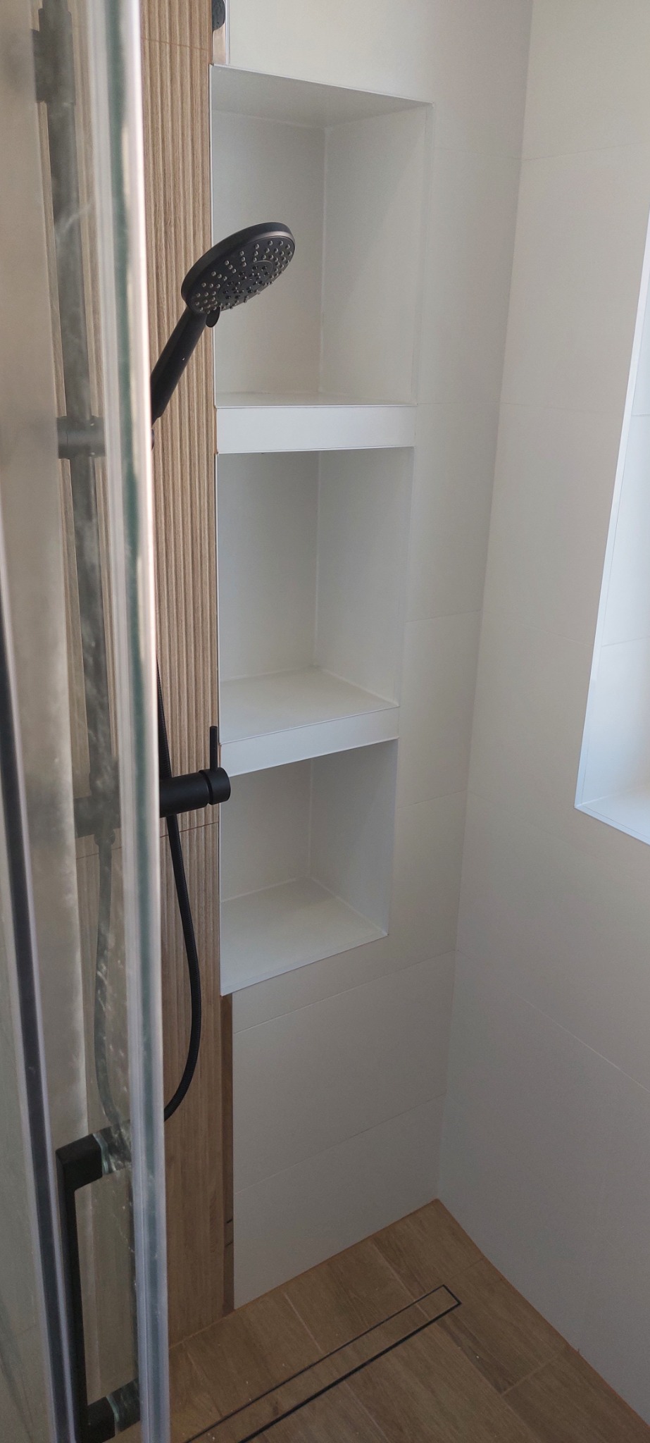 Nowoczesna kabina prysznicowa z czarną armaturą i wnękami w ścianie wyłożonymi białymi płytkami, widok na odpływ liniowy w podłodze wyłożonej płytkami imitującymi drewno.