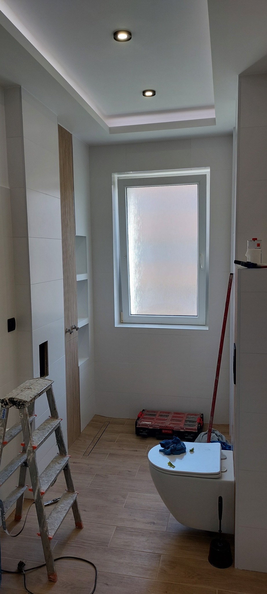 Wnętrze łazienki w trakcie prac wykończeniowych: białe płytki na ścianach, drewnopodobne płytki podłogowe, wisząca toaleta, drabina malarska, okno z matową szybą, zabudowa z półkami.