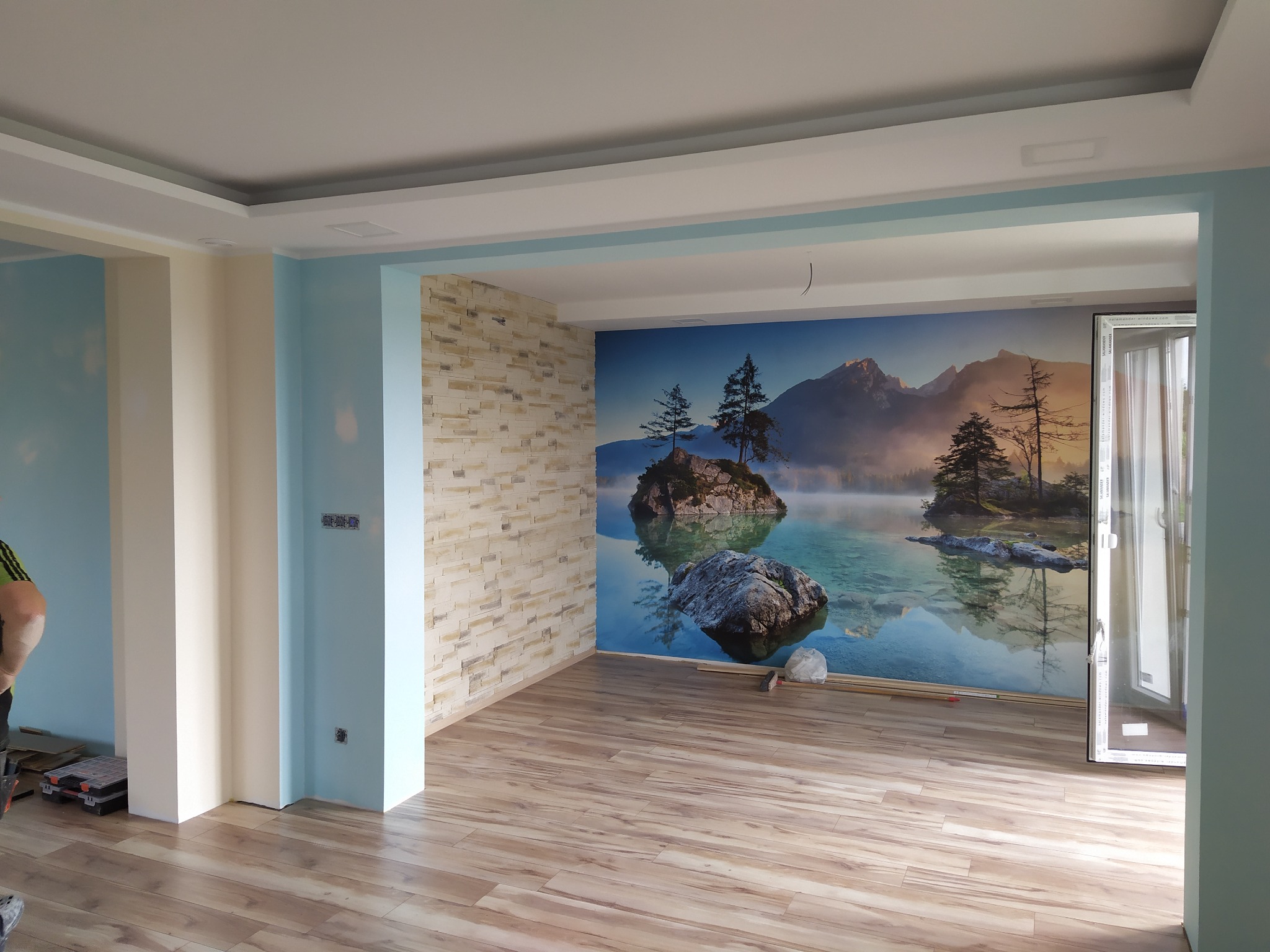 Wnętrze pokoju po remoncie z panelami podłogowymi, tapetą z motywem górskiego jeziora i ścianą wyłożoną dekoracyjną cegłą.