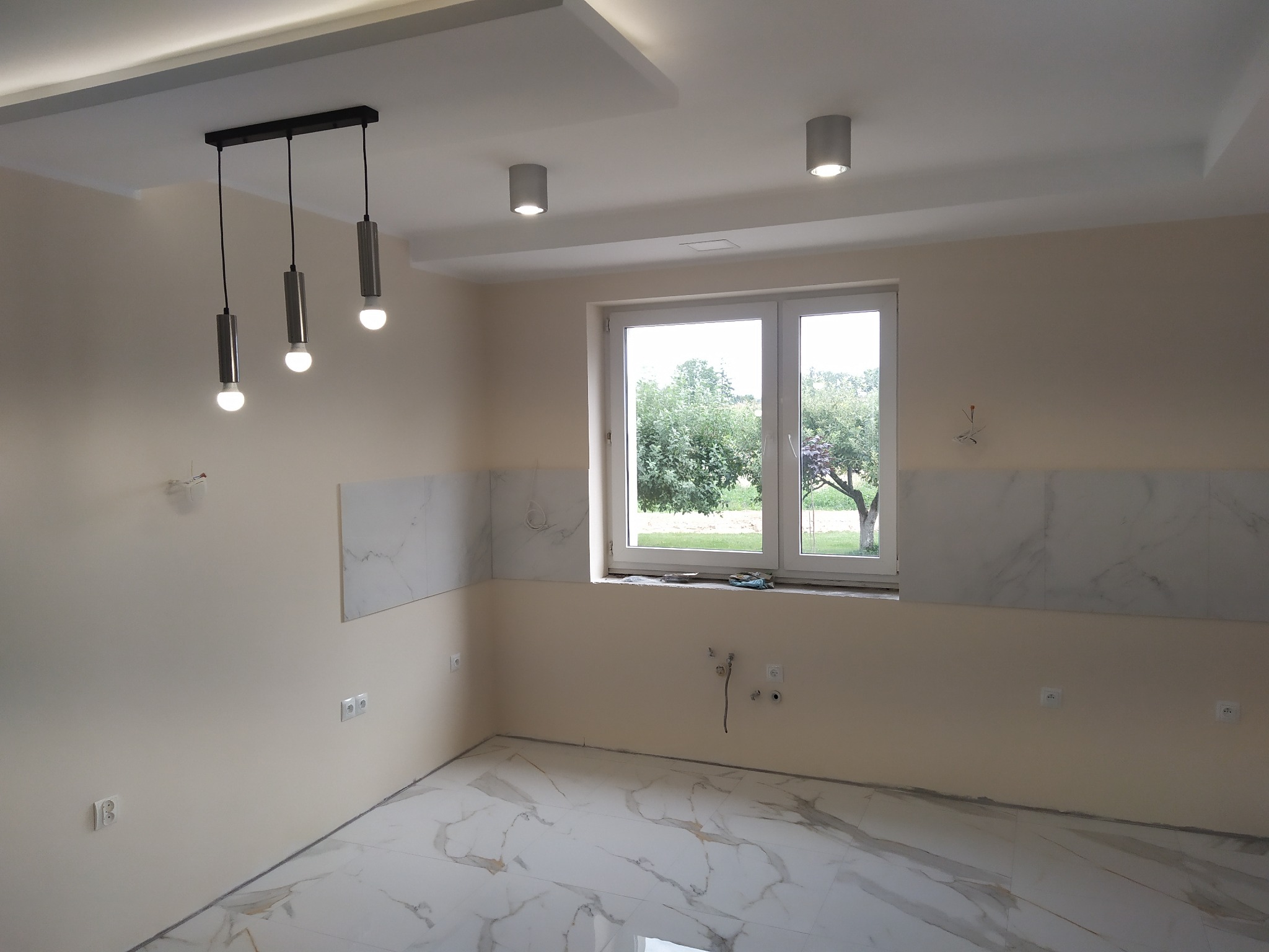 Wnętrze kuchni w trakcie prac wykończeniowych: białe marmurowe płytki częściowo ułożone na ścianie, marmurowa podłoga, okno z widokiem na zieleń, minimalistyczne oświetlenie sufitowe.