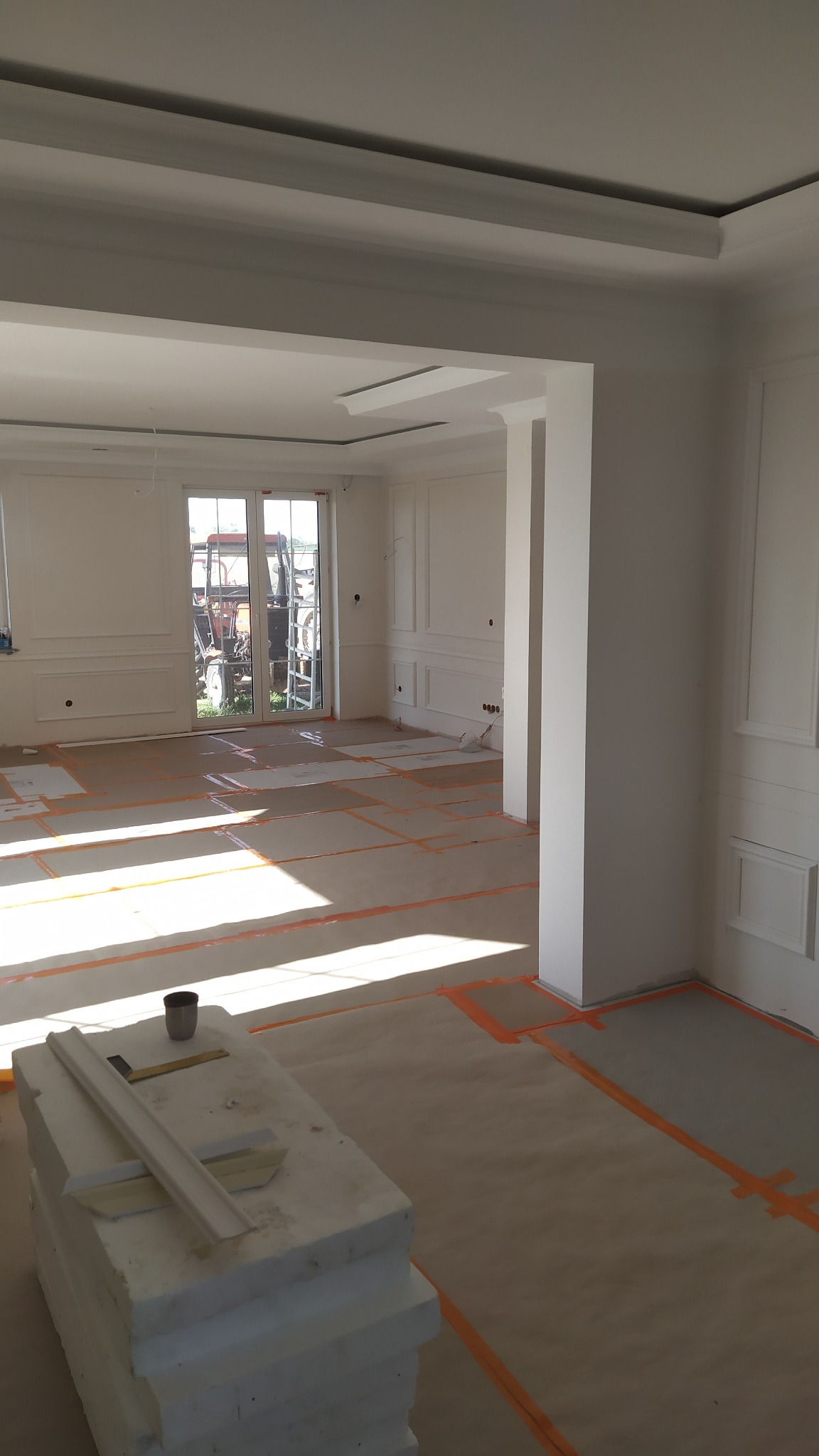 Wnętrze w trakcie prac wykończeniowych: przygotowane podłoże, sztukateria na ścianach, widoczne narzędzia i materiały, traktor za oknem.