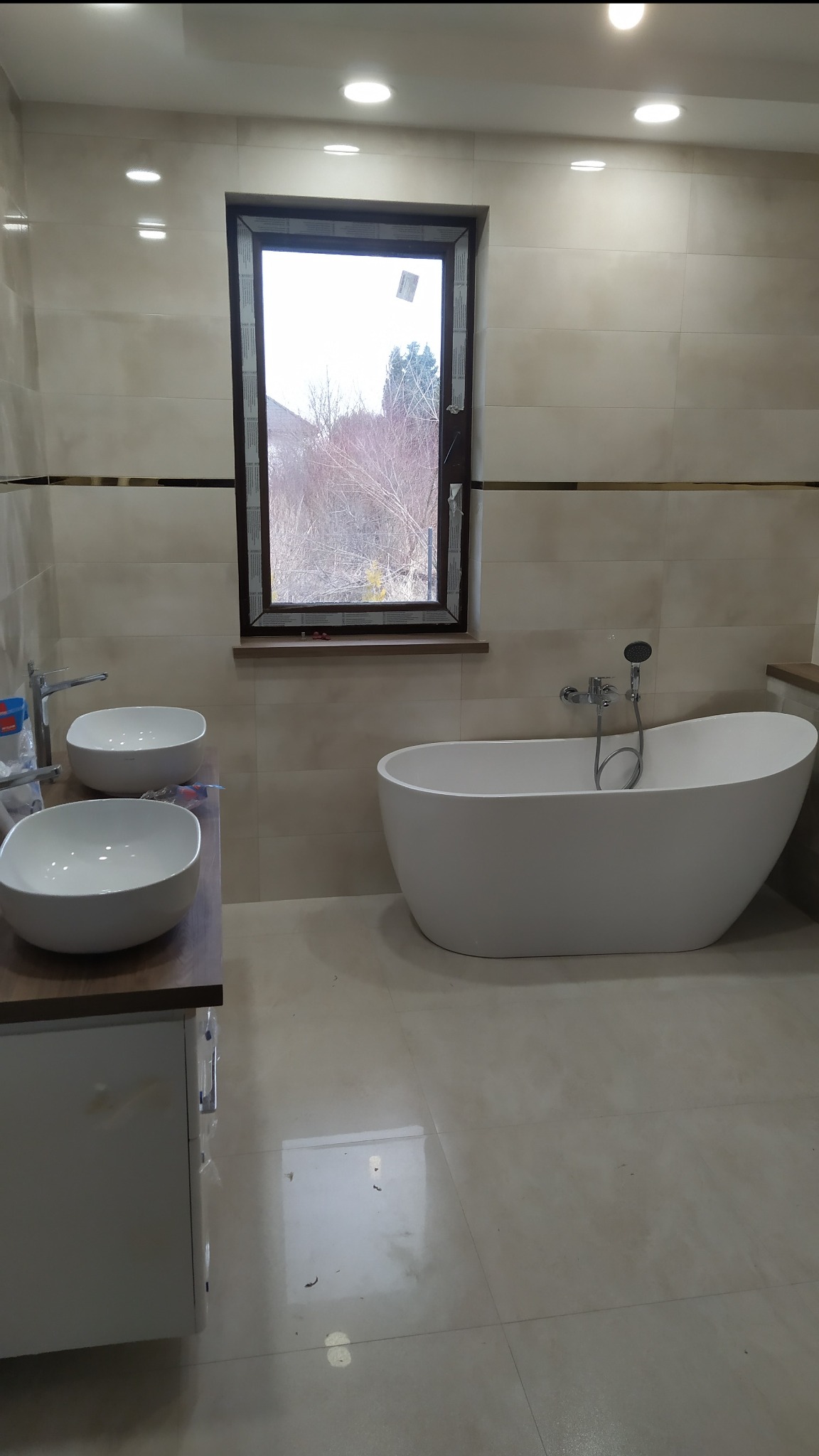 Nowoczesna łazienka z wolnostojącą białą wanną, dwiema umywalkami nablatowymi o zaokrąglonych kształtach i oknem z widokiem na drzewa bez liści.