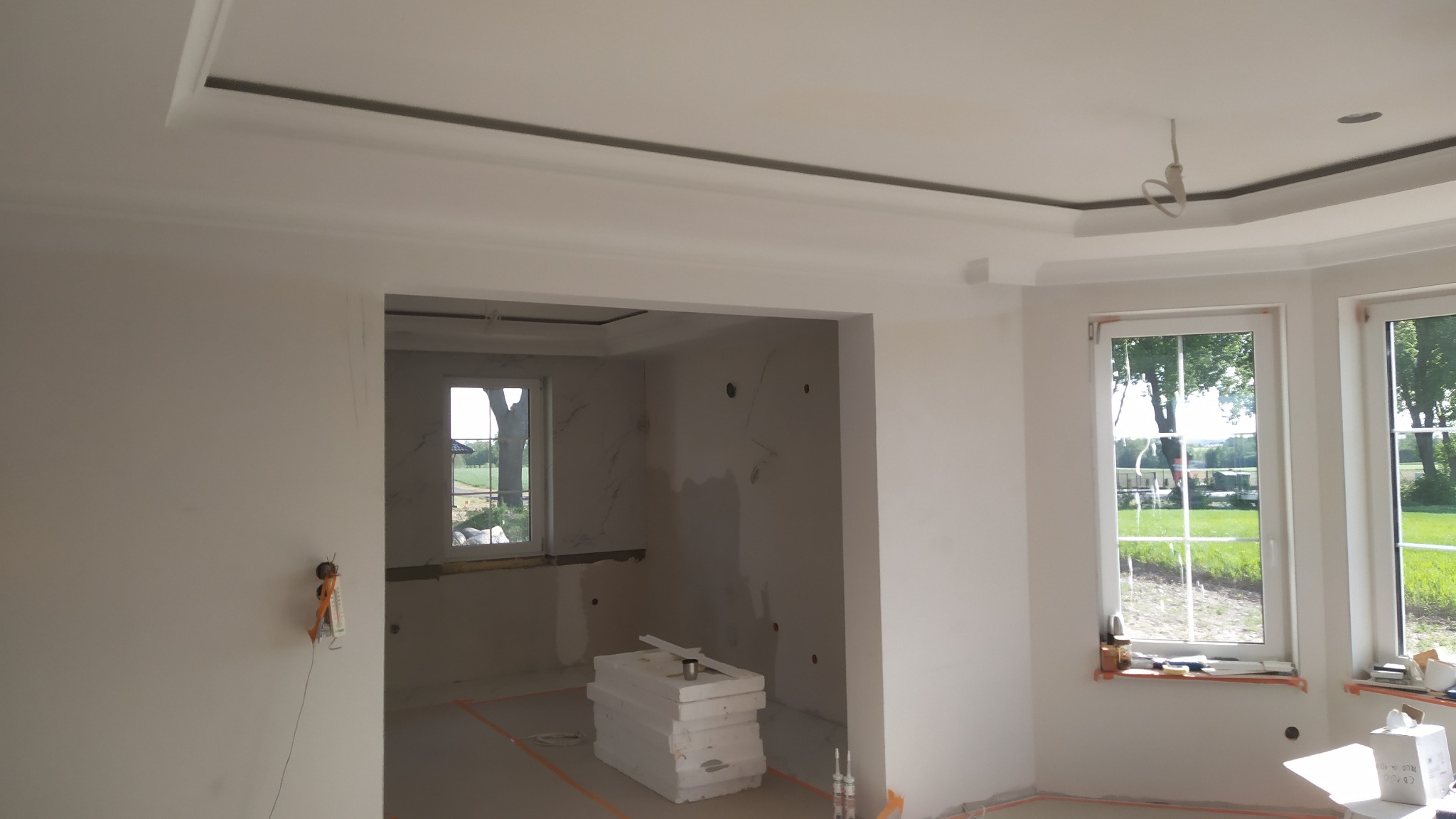 Wnętrze w trakcie prac wykończeniowych: gładkie, białe ściany, sufit z dekoracyjnymi listwami i podwieszanym elementem z oświetleniem, widoczne okna z widokiem na zieleń, materiały budowlane...