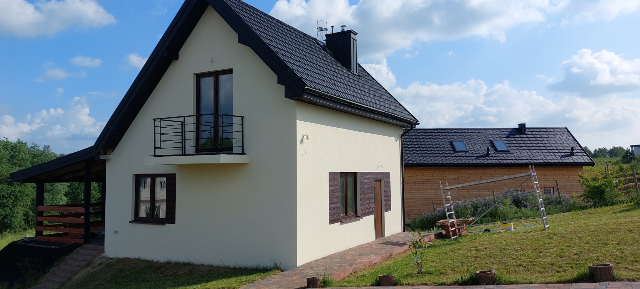 Elewacja domu jednorodzinnego w jasnym odcieniu z ciemną dachówką, widoczny balkon z metalową balustradą, okna z drewnianymi okiennicami, obok budynek gospodarczy z drewnianą elewacją, drabina...