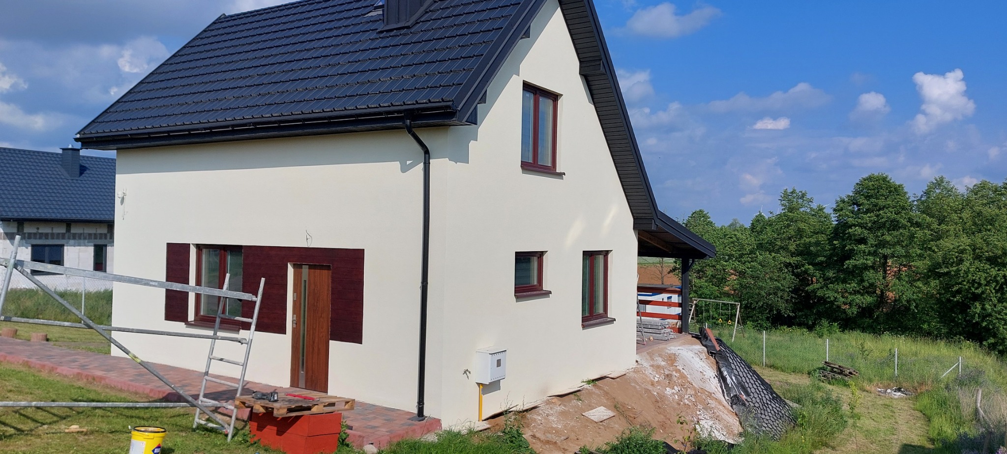 Otynkowany dom jednorodzinny z brązowymi oknami i drzwiami, widok z boku, z rozstawionym rusztowaniem i wiaderkiem farby u podstawy, na tle błękitnego nieba z chmurami.