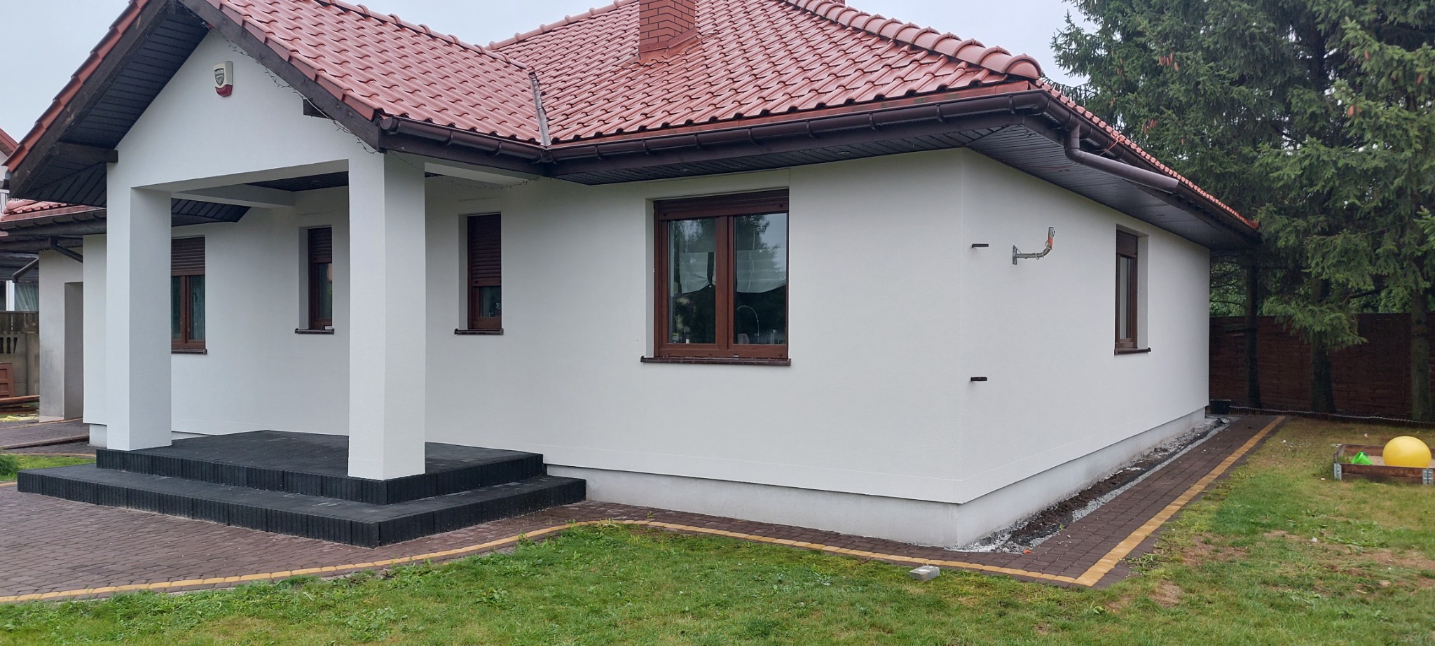 Biały dom jednorodzinny z brązową dachówką i stolarką okienną, widoczne elementy instalacji na elewacji, obok domu zielony trawnik i drzewa iglaste.