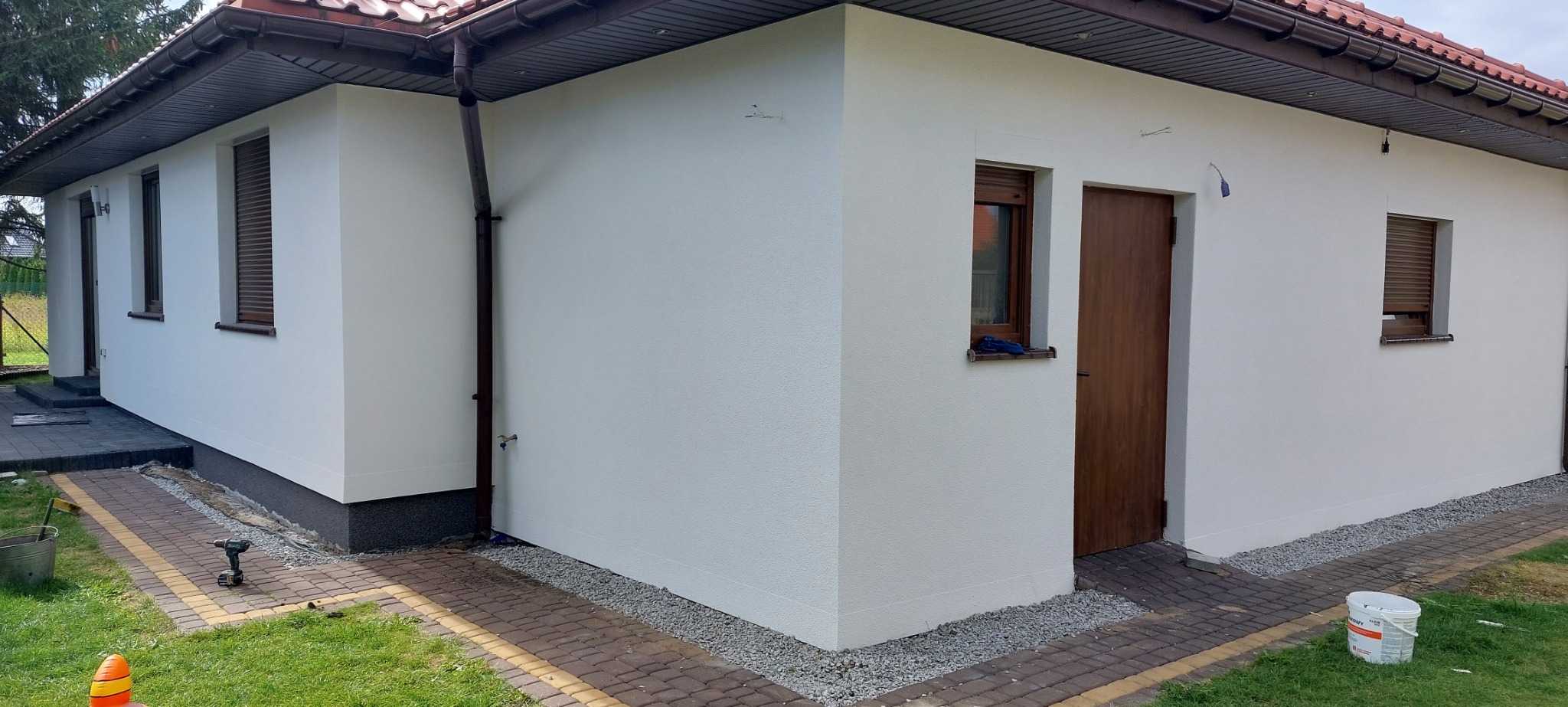 Świeżo otynkowany, biały budynek z brązowymi oknami i drzwiami, widoczne narzędzia i wiadro farby na zewnątrz, sugerujące prace wykończeniowe.