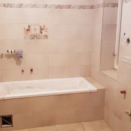 Wnętrze łazienki w trakcie prac wykończeniowych: obłożona beżowymi płytkami, z zamontowaną wanną zabezpieczoną folią, widoczne wystające rury instalacji wodnej i fragment dekoracyjnego pasa płytek...