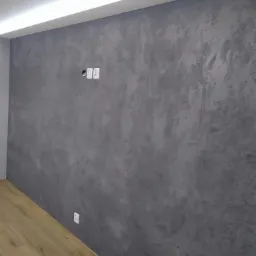 Ściana pokryta dekoracyjną szarą farbą strukturalną z widocznymi gniazdkami elektrycznymi i listwą LED podsufitową, na podłodze panele imitujące drewno.