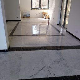 Krysbud - Wykończona podłoga z białych marmurowych płytek z czarnymi obramowaniami w przestronnym, jasnym pomieszczeniu z dużymi oknami i paletą z opakowaniami płytek w tle.