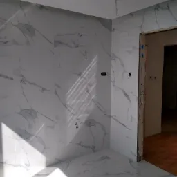 Wnętrze łazienki w trakcie wykańczania, białe płytki imitujące marmur na ścianach i podłodze, widoczne punkty instalacji elektrycznej, surowa ościeżnica drzwiowa, promienie słoneczne wpadające...