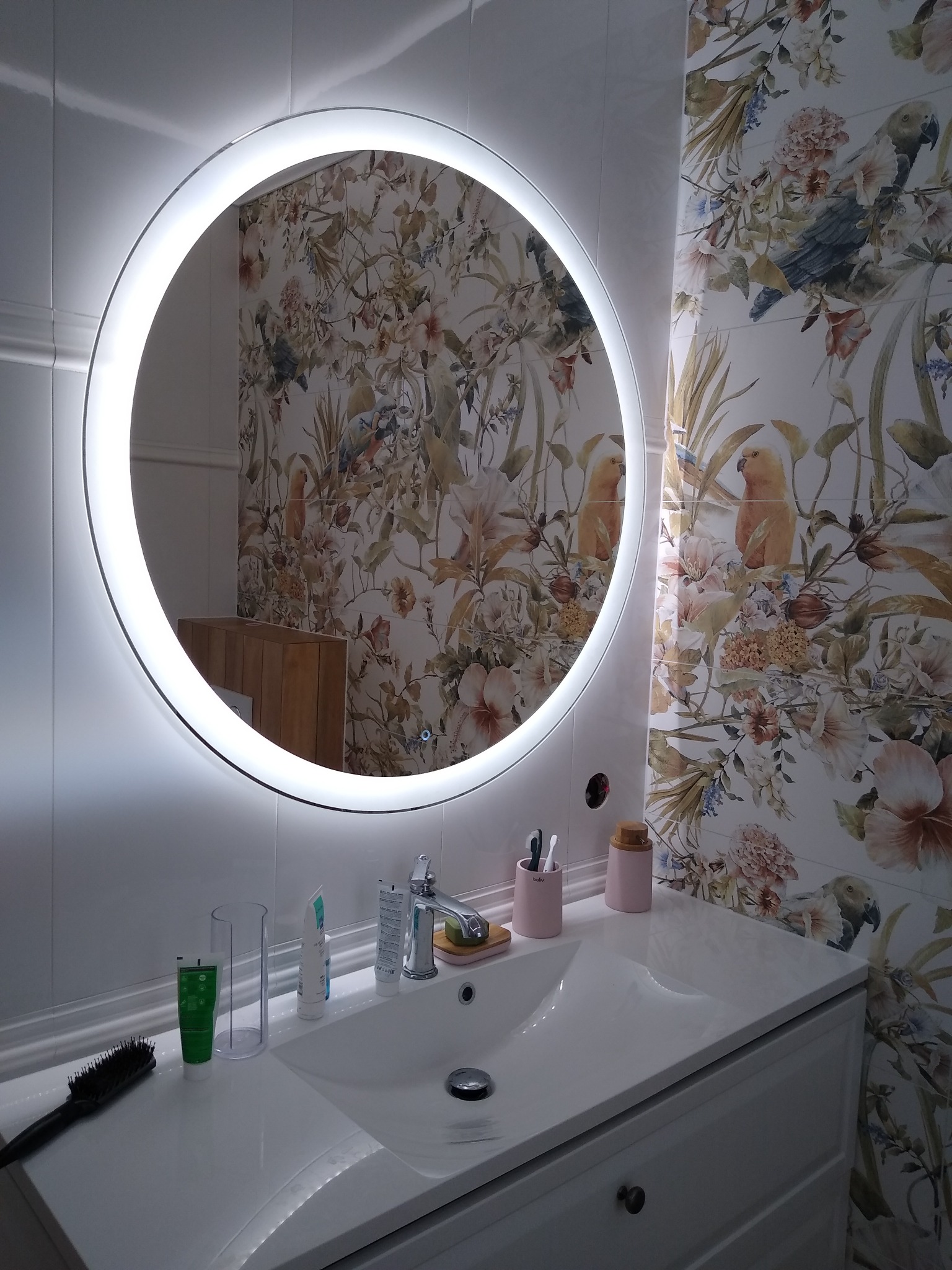Nowoczesna łazienka z okrągłym lustrem LED, umywalką w zabudowie, baterią umywalkową i płytkami z motywem kwiatowym i papugami na ścianie.