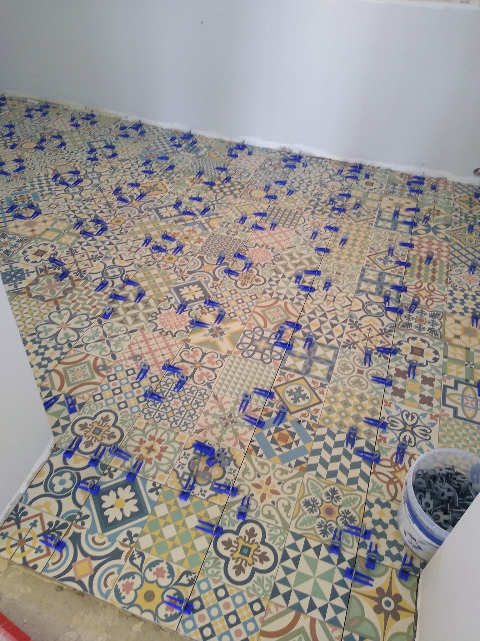 Układanie patchworkowych płytek podłogowych w trakcie prac, z użyciem niebieskich klipsów poziomujących i wiaderka z klinami przy ścianie.