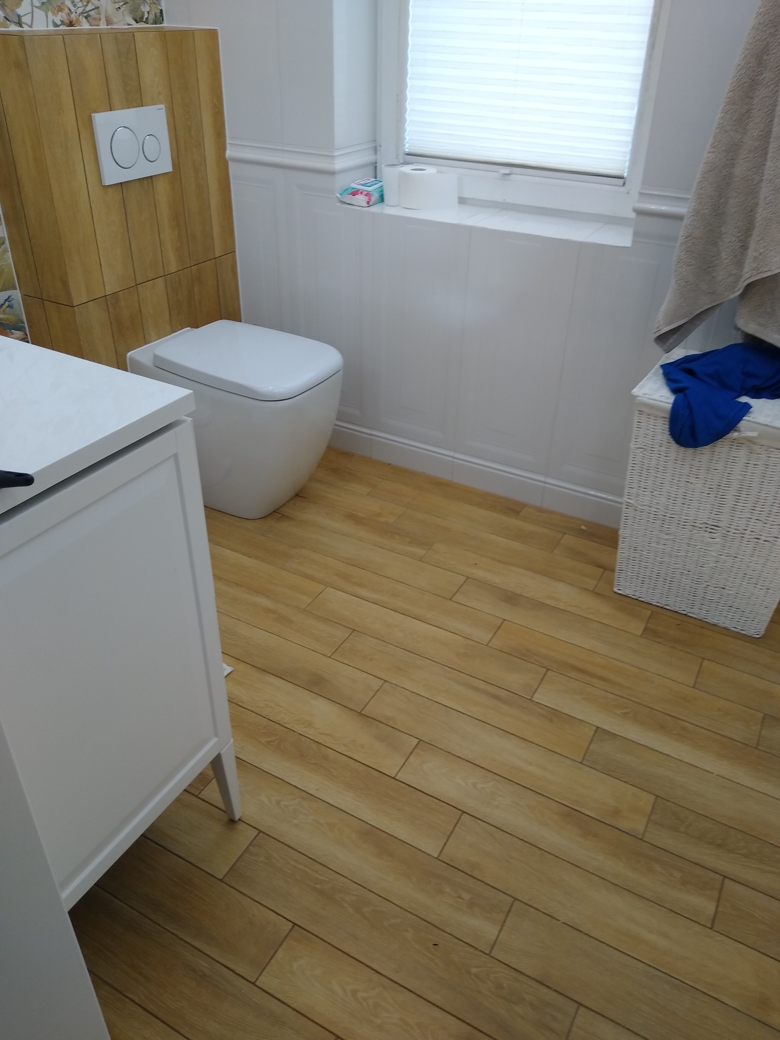 Ukończona łazienka z toaletą typu Geberit obudowaną płytkami imitującymi drewno, biała szafka pod umywalkę i podłoga wyłożona podłużnymi płytkami w kolorze drewna.