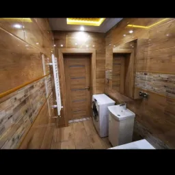 Wykończona łazienka z drewnopodobnymi płytkami na ścianach i podłodze, oświetleniem LED na suficie, grzejnikiem drabinkowym, pralką i umywalką.