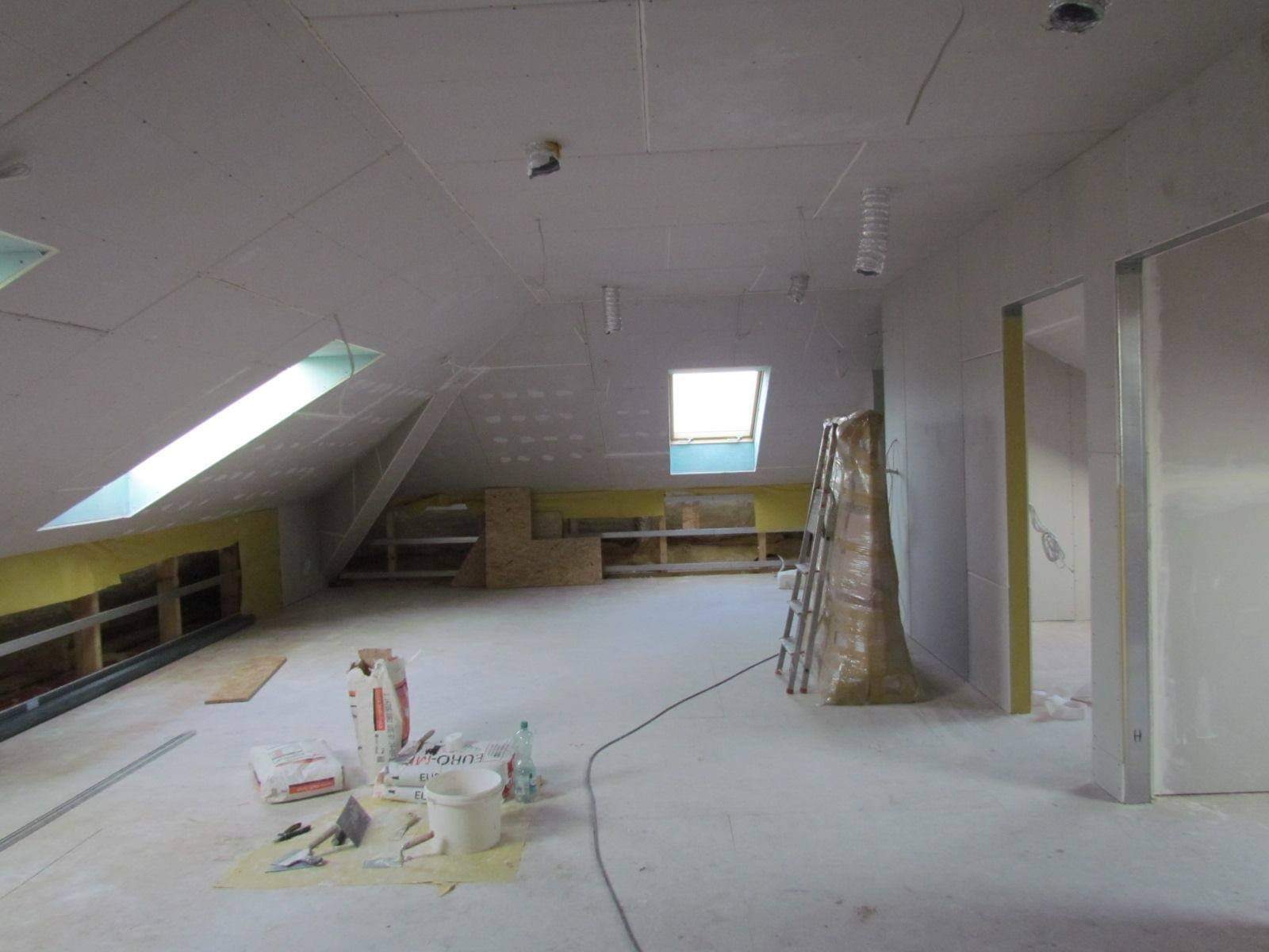 Poddasze w trakcie wykańczania: widoczne płyty karton-gips na skosach i suficie, częściowo osadzone okna dachowe, worki z zaprawą i narzędzia do szpachlowania na podłodze, drabina zabezpieczona folią.