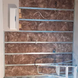 Ściana w trakcie ocieplania wełną mineralną, z widoczną instalacją elektryczną w puszce i profilem metalowym, drabina oparta o ścianę.