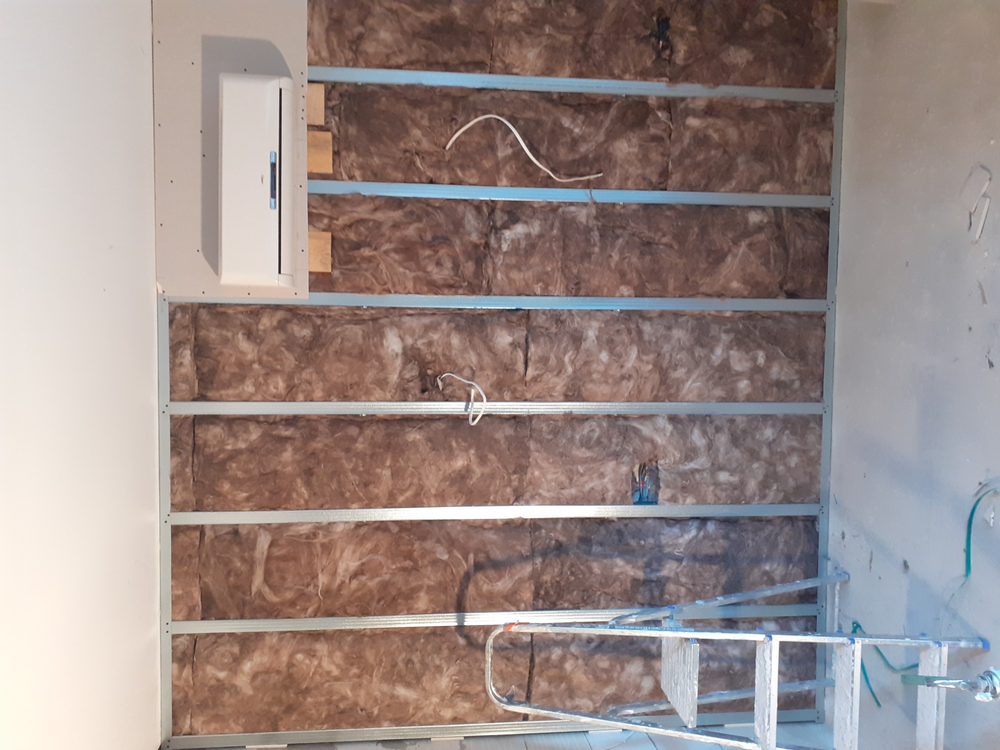 Ściana w trakcie ocieplania wełną mineralną, z widoczną instalacją elektryczną w puszce i profilem metalowym, drabina oparta o ścianę.