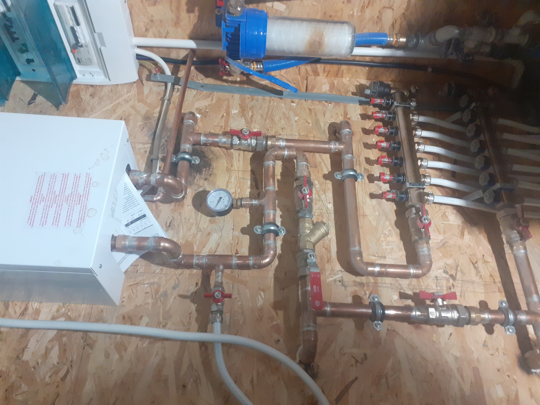 Rozbudowana instalacja hydrauliczna z miedzianych rur, zaworów, manometru i filtra wody zamontowana na ścianie z płyt OSB, widoczne liczne złączki i rozdzielacz z białymi rurami.