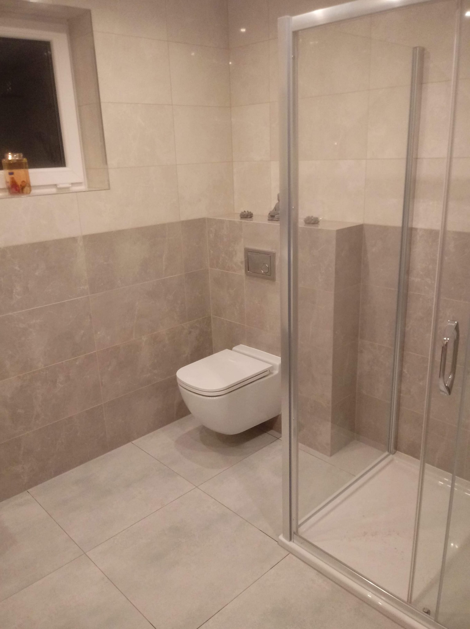 Wykończona łazienka z beżowymi płytkami, wiszącą toaletą i kabiną prysznicową z chromowanymi okuciami. Na parapecie okna stoi dekoracyjny słoik.