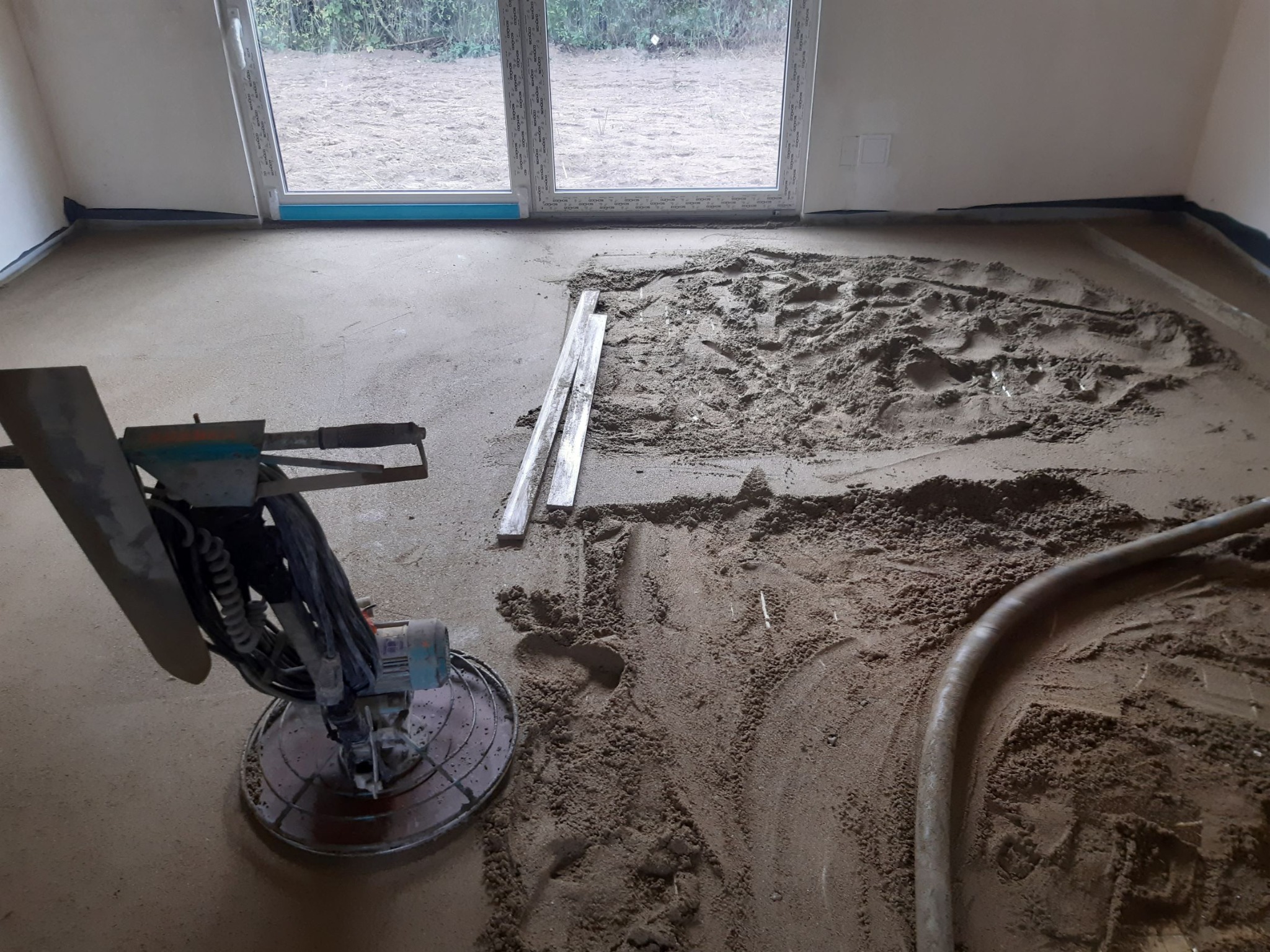Szlifowanie wylewki betonowej wewnątrz domu, widoczna zacieraczka mechaniczna, rury i deski, okno w tle.