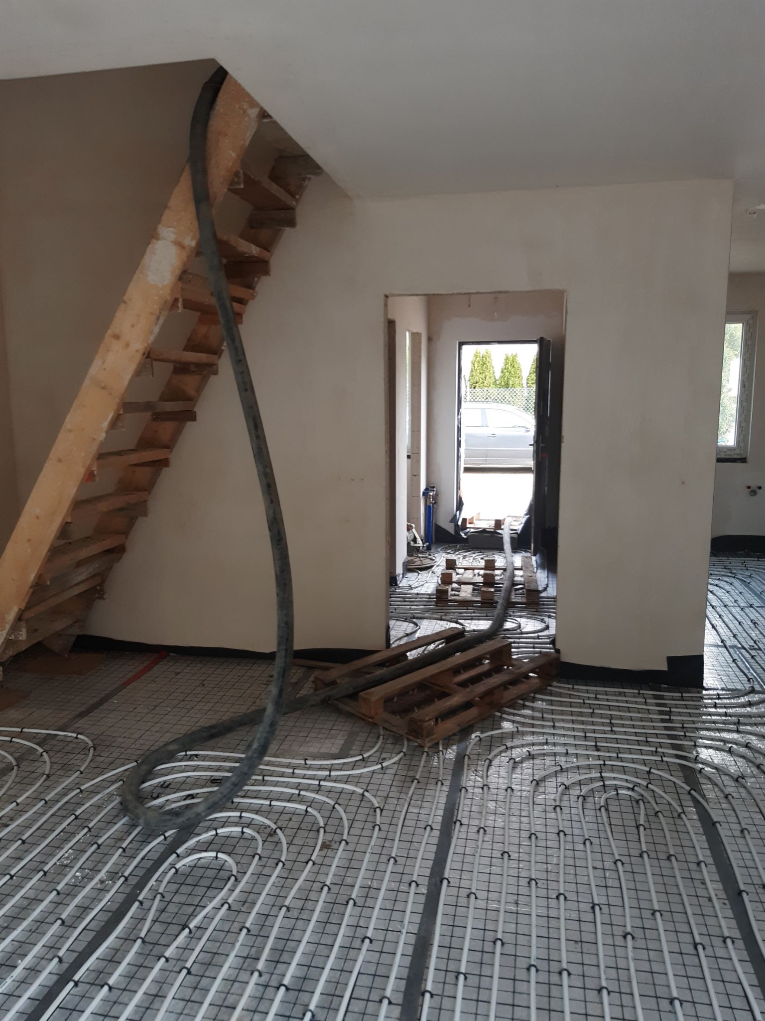 Instalacja ogrzewania podłogowego w surowym wnętrzu domu, widoczny wąż doprowadzający wylewkę, drewniane schody w tle.