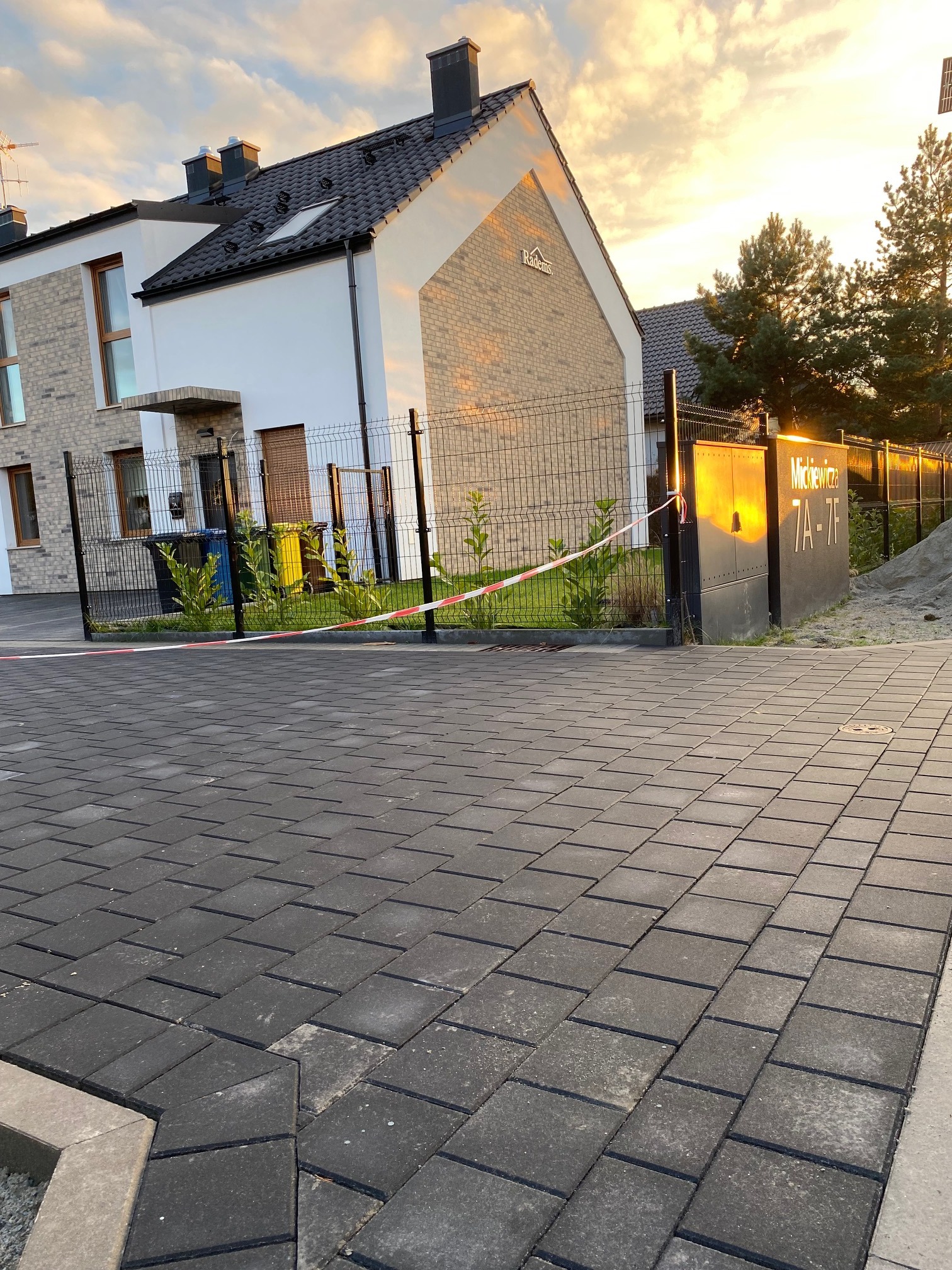 Nowoczesny dom z jasną elewacją i grafitową dachówką, widok z brukowanej drogi dojazdowej, z taśmą ostrzegawczą i tabliczką z adresem w tle, oświetlony ciepłym światłem zachodzącego słońca.