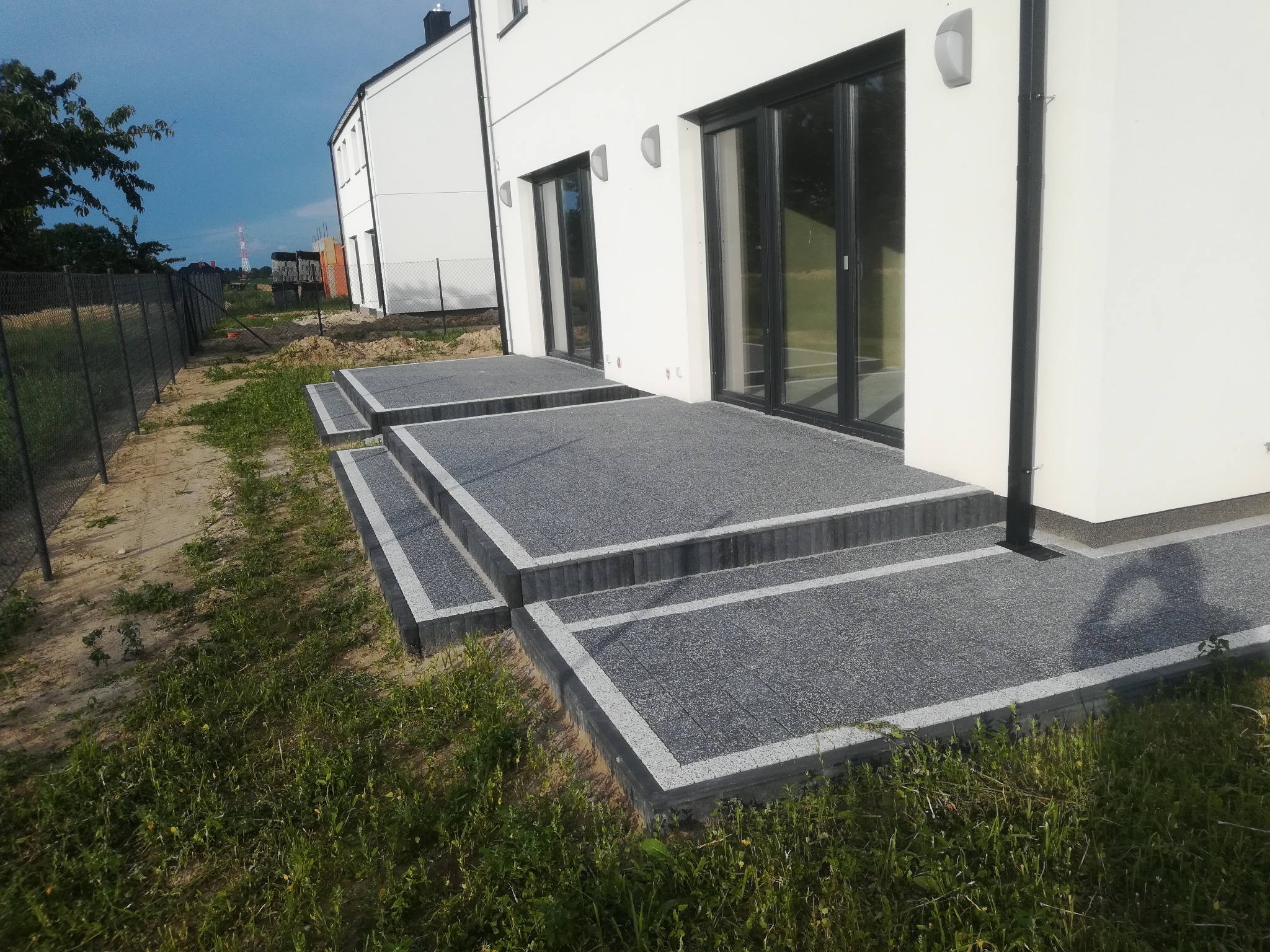 Schody wejściowe do nowoczesnego domu, wykończone szarą kostką z jasnymi obrzeżami, widok z poziomu trawnika, w tle biała elewacja i czarne ramy okienne.