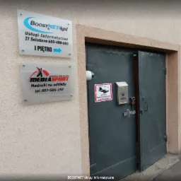 Wejście do budynku z tabliczkami firmowymi, w tym 'BoostNET.pl Usługi Informatyczne IT Solutions', kamera monitoringu nad drzwiami, skrzynka pocztowa z kłódką.
