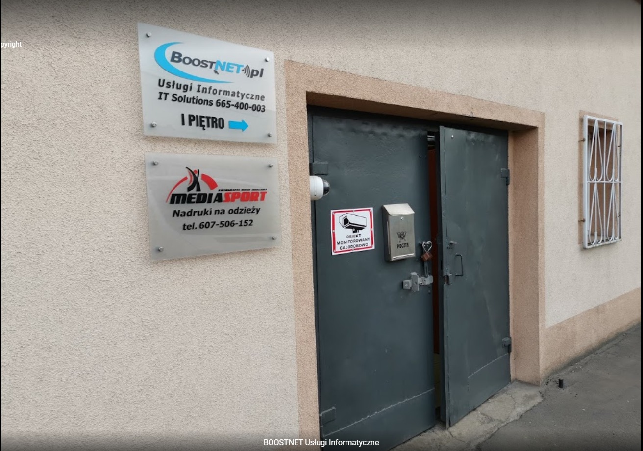 Wejście do budynku z tabliczkami firmowymi, w tym 'BoostNET.pl Usługi Informatyczne IT Solutions', kamera monitoringu nad drzwiami, skrzynka pocztowa z kłódką.