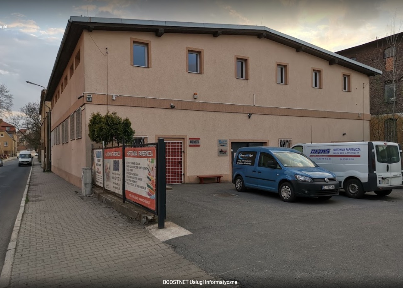 Budynek firmy z zaparkowanymi samochodami dostawczymi, w tym jeden oznaczony logo 'BoostNET Usługi Informatyczne'.