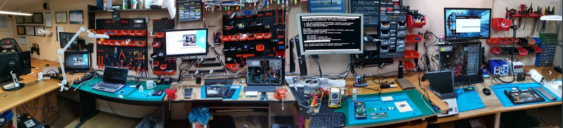 Szeroki kadr warsztatu naprawy komputerów z rozłożonymi laptopami, komputerami stacjonarnymi, narzędziami i częściami zamiennymi, z monitorami wyświetlającymi kody i oprogramowanie diagnostyczne.