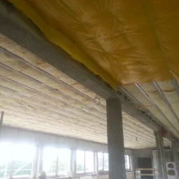 Montaż izolacji z wełny mineralnej na stropie poddasza, widoczne metalowe profile, folia paroizolacyjna i pracownik w kasku.