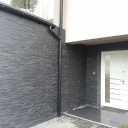 Elewacja domu wykończona szarą, łupkową okładziną kamienną, z widoczną białą, nowoczesną stolarką drzwiową i czarną rynną spustową.