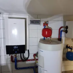 Instalacja pompy ciepła Daikin z zbiornikiem buforowym, rurami w kolorze czerwonym i niebieskim, oraz dodatkowym zbiornikiem wyrównawczym w pomieszczeniu wyłożonym białymi płytkami.