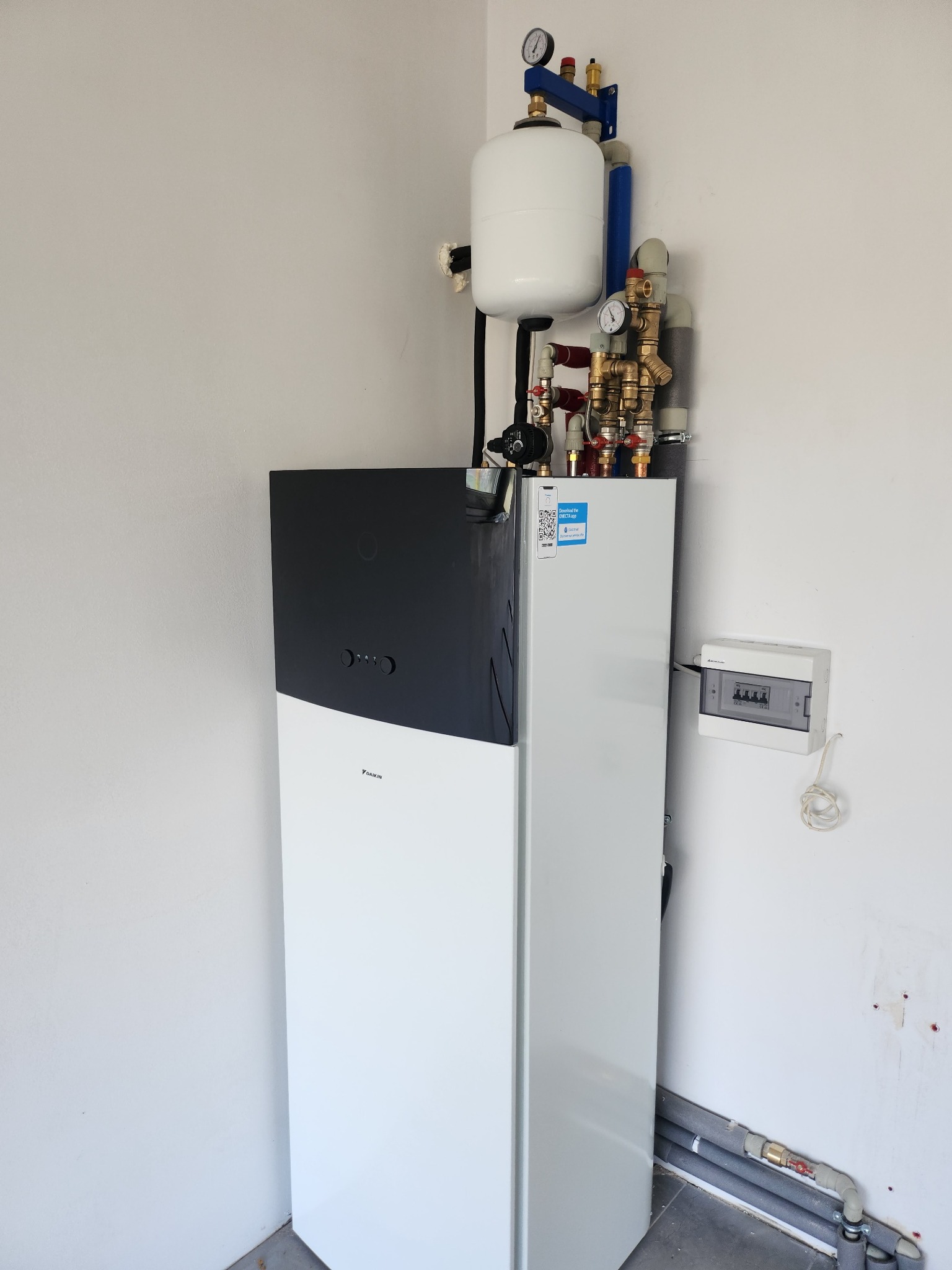 Biało-czarna pompa ciepła Daikin zamontowana w pomieszczeniu, z widocznymi rurami, manometrami i zbiornikiem wyrównawczym, obok skrzynka bezpiecznikowa na ścianie.