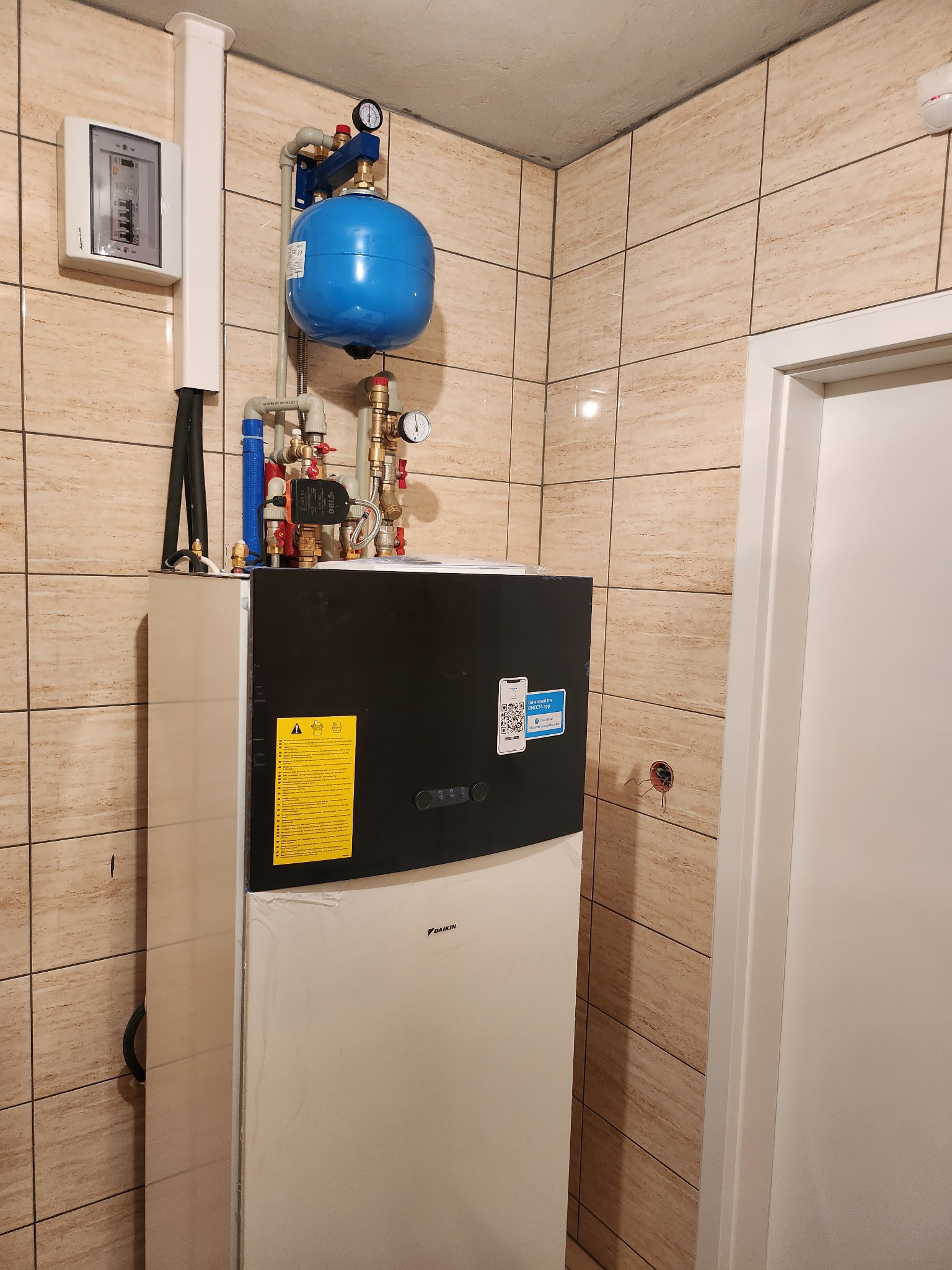 Kompaktowa pompa ciepła Daikin w pomieszczeniu z beżowymi płytkami, widoczne podłączenia hydrauliczne, manometry i panel elektryczny na ścianie.